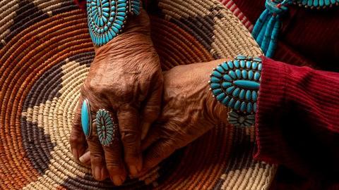 Woman's hands with two turquoise colored rings and turquoise thick bracelet on each arm folded over one another atop a woven item with star pattern