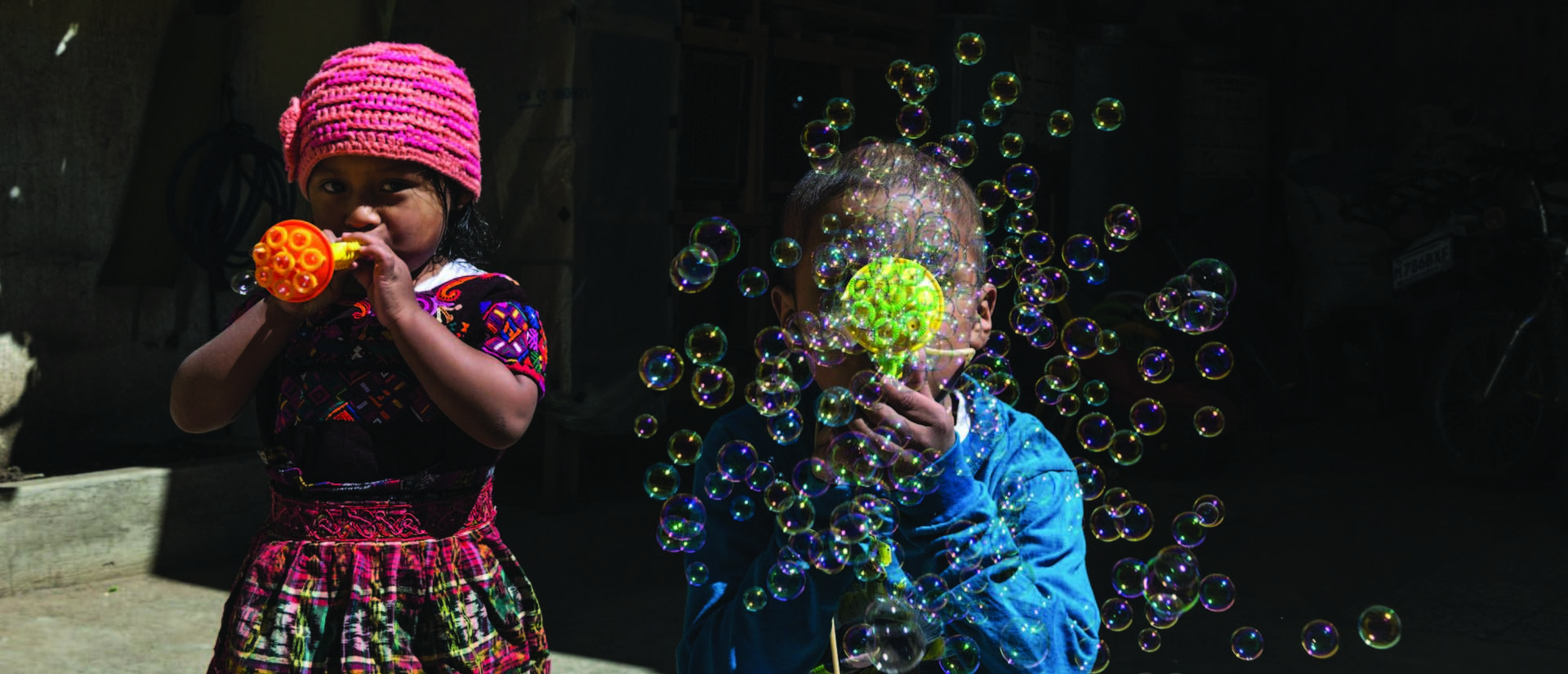 a boy in a blue shirt and a girl in a pink knit hat and print dress blowing bubbles